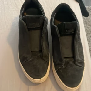 Svarta sneakers från Axel Arigato - Svarta sneakers från Axel Arigato med vit platt sula och minimalistisk design. Skorna har en stilren look utan snörning och är tillverkade i skinn. Perfekta för dig som gillar en clean och modern stil.