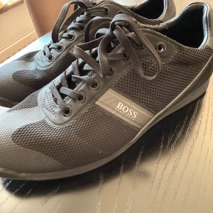 Svarta sneakers från Hugo Boss - Snygga svarta sneakers från Hugo Boss med meshpaneler och detaljer i mocka. Skorna har snörning och en diskret logga på sidan. Perfekta för dig som gillar stilrena och sportiga skor med en modern touch.