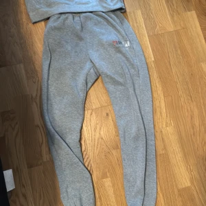 Grå mjukisbyxor från Trapstar - Säljer ett par grå mjukisbyxor från Trapstar med tryckt logga i rött, svart och vitt på ena benet. Byxorna har resår i midjan och är gjorda i mjukt bomullsmaterial, perfekta för chill dagar. Snygg och avslappnad passform.