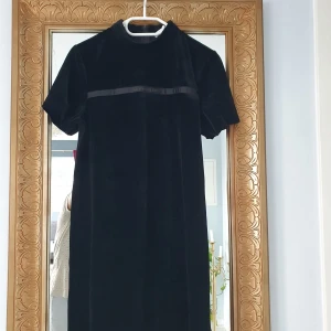 Svart sammetklänning Calvin Klein - Svart kortklänning från Calvin Klein( använd två gånger) i mjuk sammet med korta ärmar och hög hals. Klänningen har en diskret logga på ett band över bröstet och dragkedja i ryggen för enkel påklädning. Enkel och stilren design som känns lyxig och modern.