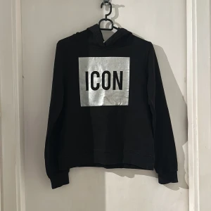 Svart hoodie med silvertryck ICON - Svart hoodie med huva och ett stort silvrigt tryck med texten ICON på bröstet. Tröjan har långa ärmar och ribbade muddar vid ärmslut och nederkant. Perfekt för dig som vill sticka ut med en cool och enkel stil.