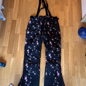 Blommiga täckbyxor från Stadium - Svarta täckbyxor med blommigt mönster från Stadium. Byxorna har hängslen, dragkedjor vid fickorna och är vattentäta med TCS Waterproof-teknik. Perfekta för vintern och håller dig torr och varm i backen.