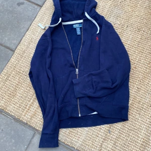 Marinblå hoodie från Polo Ralph Lauren - Marinblå hoodie från Polo Ralph Lauren med dragkedja och vit snörning i huvan. Klassisk röd logga broderad på bröstet. Tillverkad i mjuk bomullsmix med långa ärmar och känguruficka framtill. Perfekt för chill dagar eller när du vill ha en clean look.
