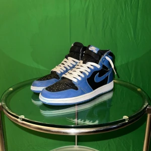 Nike Air Jordan 1 High blå/svarta - Nike Air Jordan 1 High sneakers i blått och svart med vita snören. Klassisk siluett med rund tå, perforerad tåbox och ikonisk Swoosh på sidan. Ovandel i skinn och syntet, högt skaft och vit mellansula. Perfekta för dig som vill sticka ut med en tidlös streetstyle.