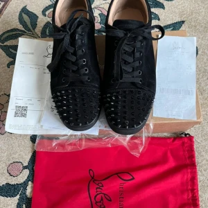 Christian Louboutin svarta sneakers - Svarta sneakers från Christian Louboutin i mocka med ikoniska röda sulor och svarta nitar på tån. Skorna har snörning och rund tå, samt en platt sula. Snygg och edgy design som sticker ut. Levereras med originalkartong och röd dustbag.