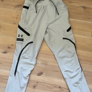 Grå träningsbyxor från Under Armour - Snygga ljusgrå träningsbyxor från Under Armour med svarta detaljer och dragkedjor på fickorna. Byxorna har smal passform, elastisk midja med snörning och är tillverkade i ett lätt syntetmaterial. Perfekta för dig som gillar sportig stil och funktionella plagg.