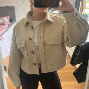 Beige overshirt med stora fickor - Säljer en beige overshirt med stora bröstfickor och snygga knappar framtill. Jackan har en croppad passform och klassisk krage, perfekt att slänga över en t-shirt. Materialet känns mjukt och har en ull-liknande look. Passar dig som gillar stilrena och trendiga plagg. Passar en xs-s 