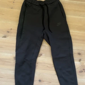 Svarta Nike tech byxor - Svarta mjukisbyxor från Nike i storlek XS. Byxorna har dragsko i midjan, smal passform och diskret Nike-logga på benet. Tillverkade i mjukt bomullsmaterial med dragkedjeficka på ena sidan. Perfekta för chill eller träning.