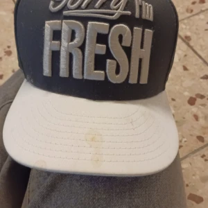 Svart och vit New Era 59FIFTY keps - Snygg svart och vit New Era 59FIFTY keps med broderad text 'Sorry I'm FRESH' framtill och 'AND YOU'RE NOT' baktill. Skärmen är vit med svart mönster undertill och insidan har en grön bandana-detalj. Perfekt för dig som vill sticka ut med en cool streetstyle.