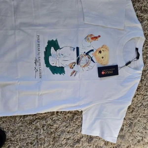 Vit Polo Bear t-shirt från Ralph Lauren - Vit t-shirt från Ralph Lauren med Polo Bear-tryck på framsidan där björnen håller tennisracketar och en dryck. Klassisk rund hals och korta ärmar. Mjuk bomullskvalitet och snygg casual vibe. Perfekt för dig som gillar sportiga och ikoniska prints.