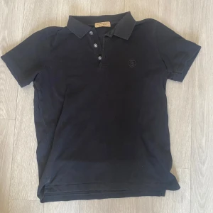 Svart Burberry pikétröja! - Klassisk svart pikétröja från Burberry med korta ärmar, krage och knappar framtill. Diskret broderad logga på bröstet och tillverkad i mjuk bomull. Perfekt för en clean och stilren look.