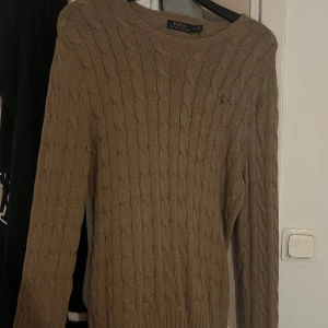Beige kabelstickad tröja Polo Ralph Lauren - Snygg beige kabelstickad tröja från Polo Ralph Lauren i 100% bomull. Klassisk rund hals och diskret broderad logga på bröstet. Perfekt för dig som gillar stilrena och tidlösa plagg med lite preppy vibe.