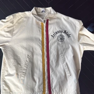 Arizona State vindjacka med tryck - Snygg beige vindjacka med Arizona State-tryck på bröstet och emblem. Jackan har dragkedja framtill med röda och gula ränder längs öppningen, två fickor och ståkrage. Perfekt för dig som gillar college-vibe och sportig stil.