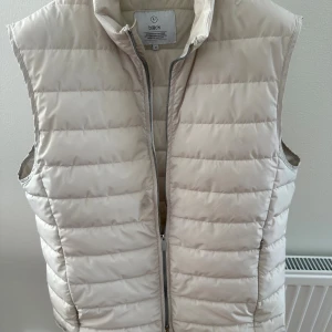 Beige dunväst från Bläck, M - Snygg krämvit dunväst från Bläck i storlek M. Västen har horisontella quiltade sömmar och en praktisk dragkedja framtill. Perfekt att slänga över en hoodie eller tröja när det är kyligt ute. Stilren och enkel design som funkar till många outfits. Har en liten fläck på framsidan men går nog bort i tvätt