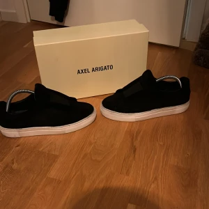Svarta sneakers från Axel Arigato - Snygga svarta sneakers från Axel Arigato med vit platt sula och minimalistisk design. Skorna har rund tå och är tillverkade i mocka med diskreta detaljer. Perfekta för dig som gillar stilrena och trendiga skor. Levereras med originalkartong och dustbag.