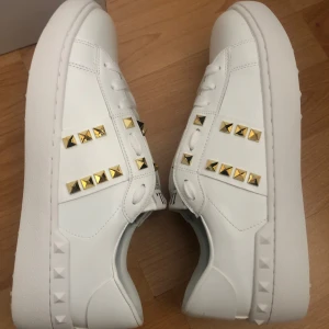 Valentino Rockstud vita sneakers - Snygga vita Valentino Garavani Rockstud sneakers med guldiga nitar och chunky sula. Skorna är i skinn och har coola pyramidformade detaljer längs sidorna och på sulan. Klassisk snörning och stilren design som sticker ut med sina metalliska accenter.