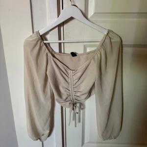 Beige croppad blus med snörning - Snygg beige blus med långa, luftiga ärmar och croppad passform. Toppen har ribbad struktur framtill och en snygg snörningsdetalj i mitten som ger en trendig look. Perfekt för dig som gillar stilrena och moderna plagg.