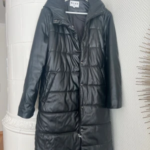 Svart lång pufferjacka från NA-KD - Lång svart pufferjacka från NA-KD med quiltad design och huva. Jackan har dragkedja och tryckknappar framtill samt två snedställda fickor. Ytan är i glansigt syntetmaterial som ger en skinnliknande look. Perfekt för kalla dagar. (En lagning i innerfodrets söm)