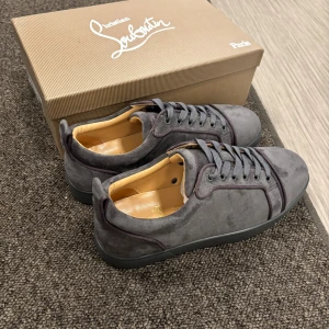 Grå mockasneakers från Louboutin - Snygga grå sneakers från Christian Louboutin i mjuk mocka med klassisk röd sula och diskreta sömdetaljer. Skorna har rund tå, platt sula och grå snörning. Perfekta för dig som vill ha en stilren men ändå lyxig look.
