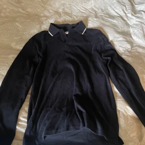 Mörkblå pikétröja från East West - Snygg mörkblå pikétröja från East West i storlek M. Tröjan har klassisk krage med vit kant och långa ärmar. Perfekt för dig som gillar en stilren och enkel look. Materialet känns mjukt och bekvämt mot huden. Har fortfarande kvar original prislapp.