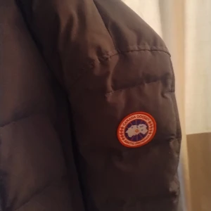 Svart dunjacka Canada Goose med päls - Säljer en svart dunjacka från Canada Goose med klassisk logga på ärmen och en fet huva med äkta päls. Jackan är rejält vadderad och har dragkedja framtill. Perfekt för kalla vinterdagar när du vill hålla stilen och värmen.