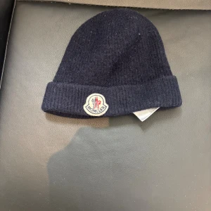 Mörkblå Moncler mössa i ull - Snygg mörkblå mössa från Moncler med klassisk uppvikt kant och broderad logotyp framtill. Tillverkad i mjuk ull som håller dig varm under kalla dagar. Perfekt accessoar för att lyfta din vinterstil.