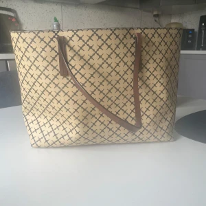 Beige Marlene Birger shoppingväska med mönster - Stor Marlene Birger shoppingväska i beige med brunt geometriskt mönster och bruna handtag. Väskan har en fyrkantig form och är tillverkad i syntetmaterial. Perfekt för dig som vill ha plats för allt du behöver under dagen.