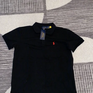 Svart pikétröja från Polo Ralph Lauren - Svart pikétröja från Polo Ralph Lauren med klassisk krage och korta ärmar. Tröjan har röd broderad logga på bröstet och är tillverkad i 100% bomull för en skön känsla. Perfekt för en clean och stilren look. Åker du på semester under de kommande kalla tiderna så är denna tröja perfekt för dig. Pris kan diskuteras vid snabbaffär!!