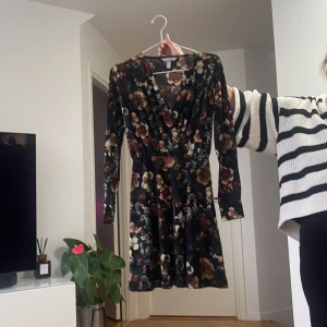 Blommig svart klänning med v-ringning - Supersnygg svart klänning med blommigt mönster i bruna och beige toner. Klänningen har v-ringning, lång ärm och markerad midja. Perfekt för dig som gillar en feminin och trendig stil.