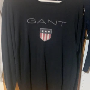 Svart långärmad tröja från GANT - Svart långärmad tröja från GANT med stort tryck av logga och sköld med stjärnor och ränder på bröstet. Klassisk rund halsringning och mjukt material som känns skönt mot huden. Perfekt för dig som gillar stilren och sportig look.