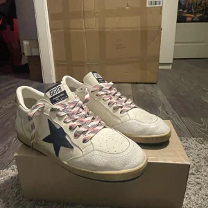 Golden goose ballstar vit/blå - Snygga GGDB Ball Star sneakers i vitt kalvskinn med en mörkblå stjärna på sidan och röda detaljer. Skorna har randiga snören i vitt, rött och blått samt svart sula undertill. Klassisk retrostil med perforerad tå och branding på plösen. Skorna är minimalt använda med endast några få creases. Dustbag och lådan följer med vid affär. 