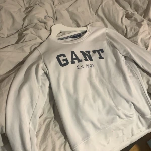 Vit sweatshirt från GANT - Vit sweatshirt från GANT med mörkblå logga och texten 'Est. 1949' på bröstet. Klassisk rund halsringning och långa ärmar. Mjuk insida och stilren design, perfekt för en chill och clean look.