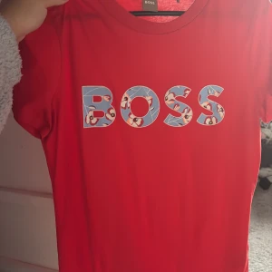 Röd BOSS t-shirt med blommigt tryck - Röd t-shirt från BOSS med coolt blommigt tryck i bokstäverna på bröstet. Klassisk rund hals och korta ärmar. Perfekt för dig som vill sticka ut med en färgstark och trendig look. Skön passform och mjukt material.
