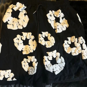 Svarta shorts med vit mönster, strl S - Coola svarta shorts med stora vita mönster som ser ut som blommor eller cirklar. De har resår i midjan och snörning för extra passform. Perfekta för sommaren och tillverkade i mjuk bomull som känns skön mot huden.