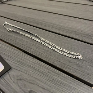 Silverfärgad kedjehalsband - Stilrent halsband i silverfärg med platt kedjedesign. Kedjan har en klassisk look och passar perfekt till streetwear eller för att lyfta en enkel outfit. Längden är medellång och halsbandet har ett smidigt lås. Perfekt accessoar för dig som gillar minimalistisk stil.