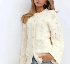 Vit stickad kofta gina - Mysig vit stickad kofta med grovt flätmönster och rund hals. Koftan har långa ärmar och stängs med knappar framtill. Perfekt att slänga över en t-shirt för en chill look.