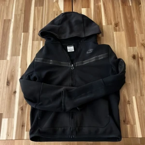 Svart Nike hoodie med dragkedja - Svart tech hoodie från Nike med hel dragkedja och luva. Tröjan har vit Nike-logga på bröstet och en stilren design med svarta detaljer över bröstet. Perfekt för chill, träning och för hösten 🍂med långa ärmar och bekväm passform 