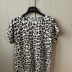 Leopardmönstrad t-shirt New Collection - Cool t-shirt med leopardmönster från New Collection. T-shirten är vit i grunden med svarta och beige fläckar, har rund hals och korta ärmar. Perfekt för dig som vill sticka ut med en trendig och djurinspirerad look.