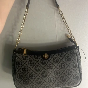 Svart och grå axelväska från River Island - Snygg axelväska från River Island i svart och grått med monogrammönster. Väskan har guldfärgade kedjedetaljer på axelremmen och dragkedja upptill. Perfekt storlek för det viktigaste och en stilren accessoar till din outfit.