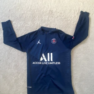 Mörkblå PSG långärmad fotbollströja - Snygg mörkblå långärmad PSG fotbollströja från Jordan med dragkedja vid halsen. Tröjan har vita tryck med klubbmärke, sponsorloggor och diskreta ränder i materialet. Röd, vit och blå detalj på ärmen. Perfekt för dig som älskar fotboll och streetwear.