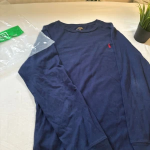 Mörkblå långärmad tröja Polo Ralph Lauren - Säljer en mörkblå långärmad tröja från Polo Ralph Lauren i 100% bomull. Klassisk design med rund hals och den ikoniska röda loggan broderad på bröstet. Perfekt för en clean och stilren look.