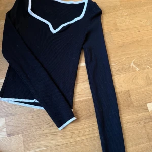 Svart ribbad långärmad topp med vit kant - Svart ribbad långärmad topp med vit kant längs den v-ringade halsen och vid ärmslut och nederkant. Snygg och enkel modell med figurnära passform, perfekt till jeans eller kjol.