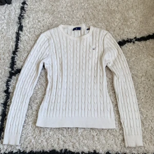 Vit kabelstickad tröja från Gant - Säljer en vit kabelstickad tröja från Gant med rund halsringning och logga broderad på bröstet. Tröjan har lång ärm och ribbade muddar vid ärmslut och nederkant. Säljer då den inte kommer till användning, säljer endast via instabox 