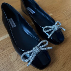 Svarta ballerinaskor Steve Madden rosett - Svarta ballerinaskor från Steve Madden i glansigt material med fyrkantig tå. Dekorerade med en stor glittrig rosett framtill som ger extra bling. Platt sula och klassisk ballerinaform, perfekta för att lyfta vilken outfit som helst. Helt nya och oanvända. Storlek 40. Nypris 1319kr.