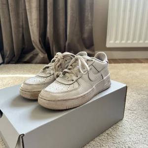 Nike Air Force 1 vita sneakers - Klassiska Nike Air Force 1 sneakers i helvitt skinn med perforerad tå och snörning. Snygg och tidlös modell med låg profil och rund tå, perfekt till streetwear. Swoosh-logga på sidorna och bekväm platt sula.