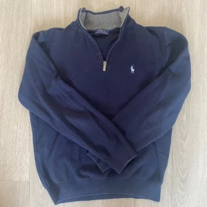 Marinblå half zip tröja Polo Ralph Lauren - Snygg marinblå tröja från Polo Ralph Lauren med halv dragkedja och broderad logga på bröstet. Tröjan har lång ärm, ribbade muddar och en mjuk insida i grått. Perfekt för lager-på-lager och chill stil.