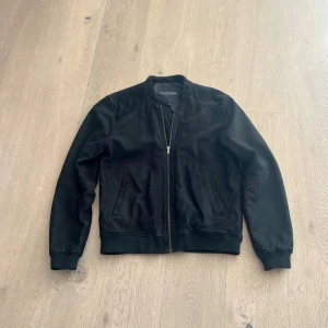 Svart bomberjacka i äkta mocka från Bruun & Stengade - Snygg svart bomberjacka från Bruun & Stengade i 100% äkta mocka med foder i polyester. Jackan har dragkedja framtill, ribbade muddar och två snedställda fickor. Perfekt för dig som vill ha en stilren och klassisk look.