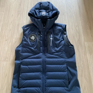 Blå dunväst från Sail Racing XS - Säljer en blå dunväst från Sail Racing i storlek XS. Västen har huva, dragkedja framtill, två sidofickor med dragkedja och coola loggor på bröstet. Perfekt för dig som gillar sportig och funktionell stil.