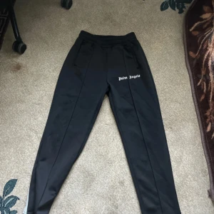 Svarta mjukisbyxor från Palm Angels - Svarta mjukisbyxor från Palm Angels med vit logga på vänster lår. Byxorna har elastisk midja och raka ben med en sportig look. Tillverkade i ett glansigt syntetmaterial som ger en clean och modern vibe.