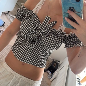Svartvit rutig croppad blus med volang - Superfin croppad blus i svartvitt rutigt mönster med volangdetaljer och stor rosett framtill. Offshoulder-modell med korta ärmar och söt, feminin vibe. Perfekt för dig som gillar trendiga och lekfulla toppar.
